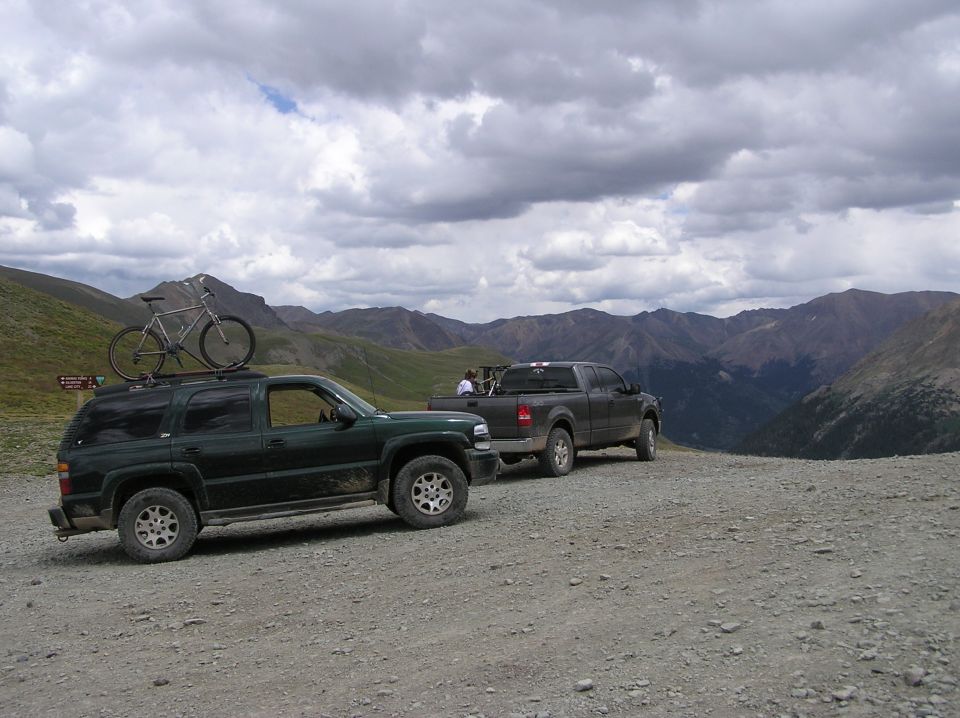 Dark green SUV with bicycle on roof rack, parked on a gravel road with a pickup truck behind it