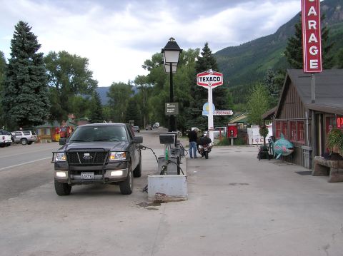 1986 Ford Bronco - A Ford F-150 pickup truck is parked at a gas pump in a rural town with mountains in the background.