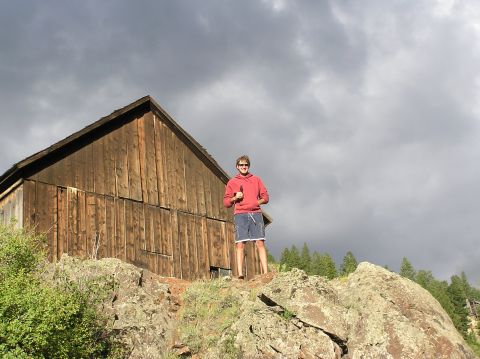 1986 Ford Bronco - Man with a bottle stands on rocks in front of a wooden building with a stormy sky overhead.