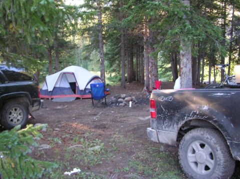 1986 Ford Bronco - A campsite scene in a forest with a tent, chairs, campfire, and two pickup trucks.