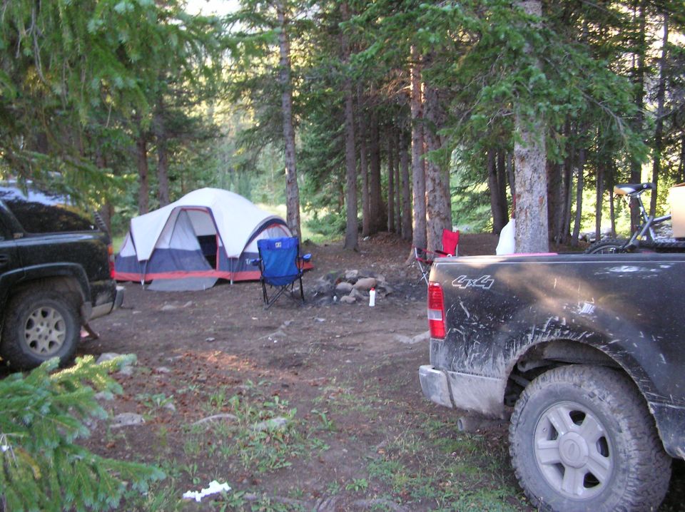 Campsite in woods with a tent, campfire, and two pickup trucks parked nearby