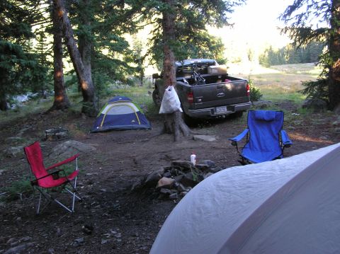 1986 Ford Bronco - A campsite is set up in a wooded area with a pickup truck, tents, and chairs around a fire pit.