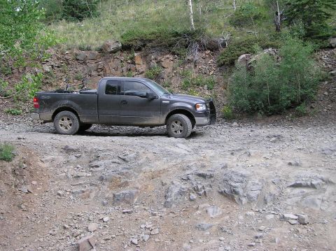 1986 Ford Bronco - A four-door pickup truck navigates a rough, off-road trail with rocky terrain and sparse vegetation.