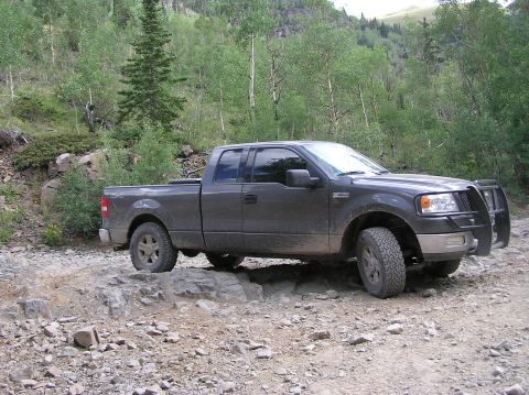 1986 Ford Bronco - A dark gray Ford F-150 navigates a challenging rocky path in a wooded mountain area.