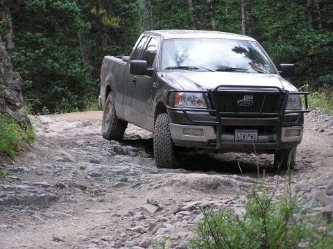 1986 Ford Bronco - A dark gray Ford F-150 navigates a challenging, rocky off-road trail in a wooded area.