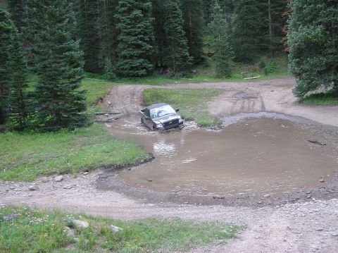 1986 Ford Bronco - A dark gray Ford F-150 navigates a muddy obstacle on a forest road.