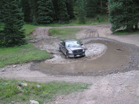 1986 Ford Bronco - A dark pickup truck tackles a muddy obstacle on a winding dirt road surrounded by trees.