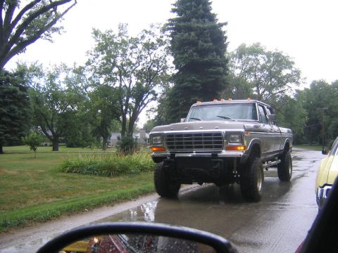 1978 Ford Bronco - A lifted 1970s Ford F-Series truck drives down a wet road, partially obscured by a yellow vehicle.