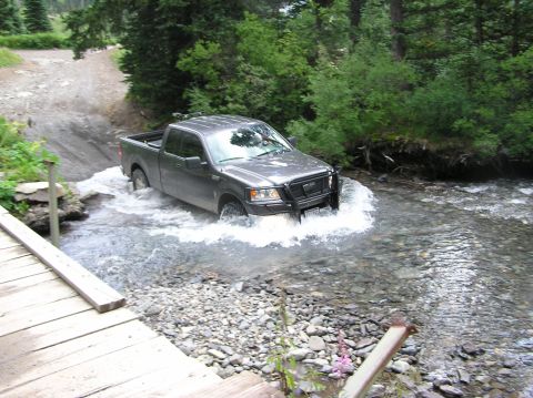1986 Ford Bronco - A gray pickup truck navigates a shallow stream, its front grille suggesting a 2004 model year.