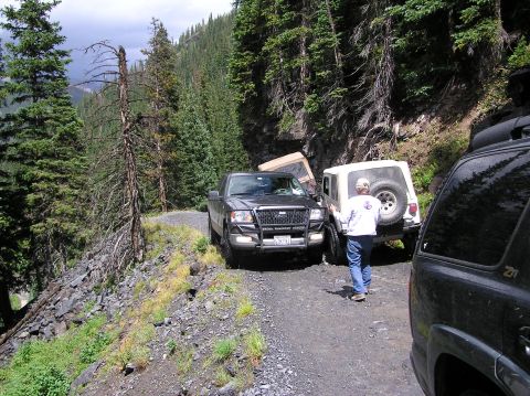1986 Ford Bronco - Several off-road vehicles navigate a narrow mountain path with dense trees on either side.