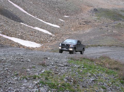 1986 Ford Bronco - A dark pickup truck navigates a gravel road in a , mountainous landscape with snow patches on the slopes.