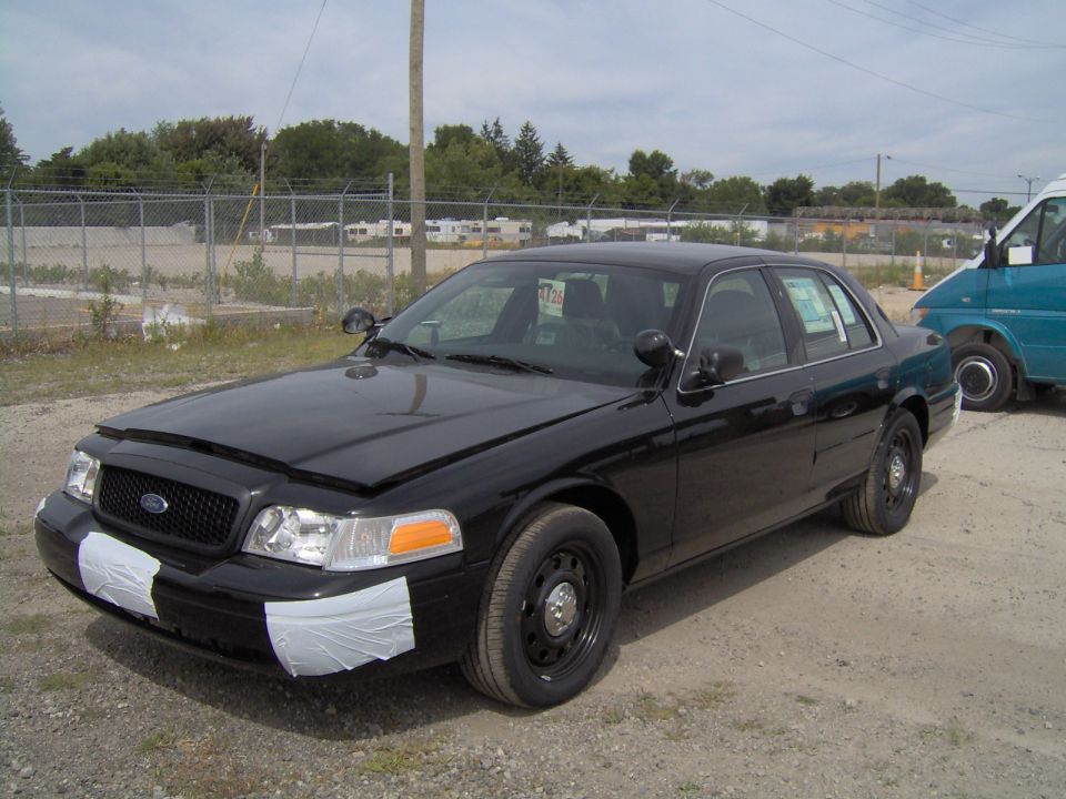 Black Ford Crown Victoria sedan, front 3/4 view, with hood bra and taped bumper on gravel lot.