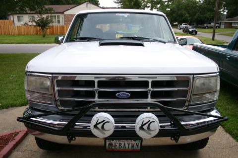 1994 Ford Bronco - Front view of a white 1990s Ford Bronco parked on a driveway, showing its chrome grille with blue Ford emblem, dual headlights, a black tub…