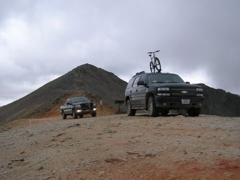 1986 Ford Bronco - Two vehicles, an SUV with a bike rack and a pickup truck, are parked on a dirt road with a mountain in the background.