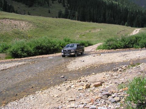 1986 Ford Bronco - A dark gray Ford F-150 pickup truck navigates a rocky stream in a mountainous, wooded area.