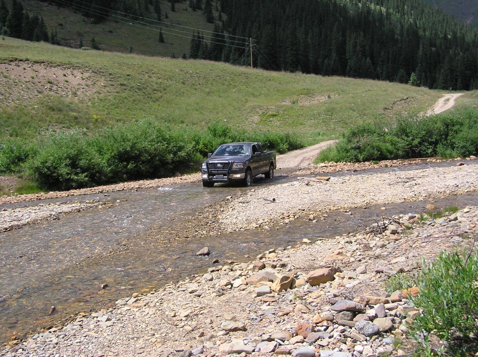 Dark gray Ford F-150 pickup truck driving through a shallow, rocky stream bed