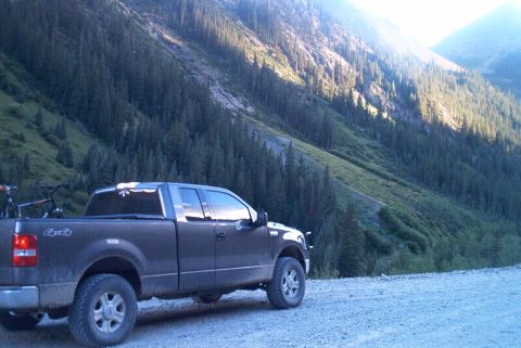 1986 Ford Bronco - A dark-colored Ford F-150 pickup truck is parked on a gravel road, with a steep, forested mountainside behind it.