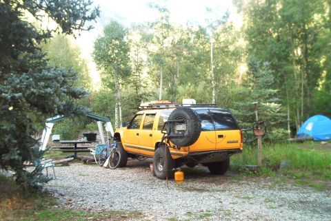 1986 Ford Bronco - A yellow SUV is parked at a campsite with a spare tire mounted on the back, next to a picnic table and a bicycle.
