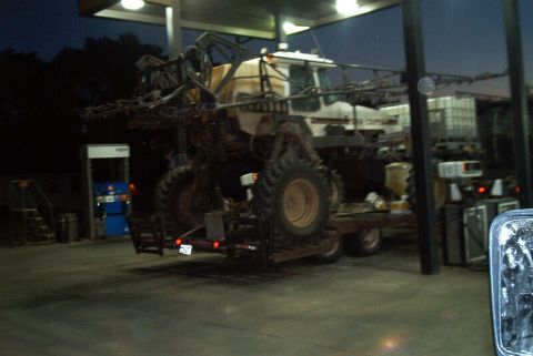 1986 Ford Bronco - Large agricultural sprayer truck being towed on a trailer at a gas station at dusk.