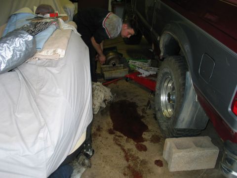 1995 Ford Bronco - Mechanic works on the transmission of a maroon and grey SUV, with fluid spilled on the garage floor.