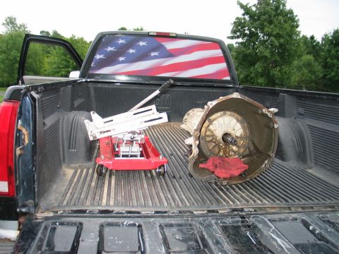 1995 Ford Bronco - A transmission and a red jack are in the bed of a pickup truck with an American flag decal on the rear window.