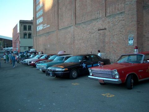2001 Ford Crown Victoria - A line of cars, including a black police car and a red emergency vehicle, are parked outside a brick building.