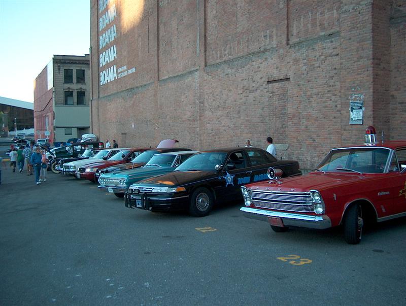 Row of police cars at a show beside a brick warehouse, red 1960s Ford in front
