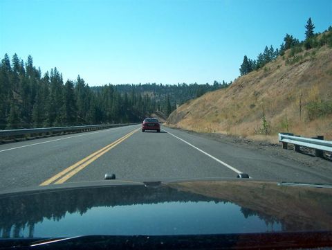 2001 Ford Crown Victoria - A red sedan travels down a highway surrounded by trees and a dry, grassy hillside under a clear blue sky.
