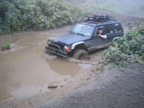 1992 Ford Bronco - A Jeep Cherokee XJ navigates a muddy off-road obstacle, with its front right wheel deep in the mire.