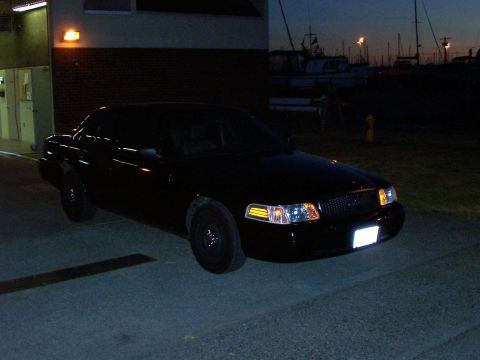 2003 Ford Crown Victoria - A black 2003 Ford Crown Victoria is parked near a building at dusk, with boats visible in the background.