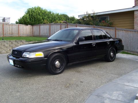 2003 Ford Crown Victoria - A black sedan is parked on a paved driveway in front of a wooden fence and a house.