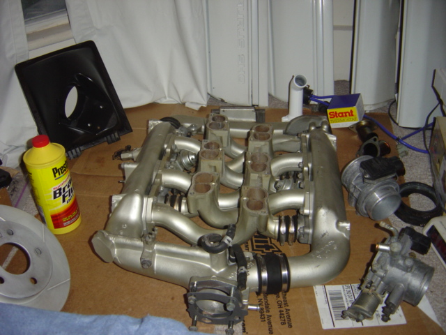 Silver engine intake manifold with multiple pipes and throttle bodies, next to a brake rotor and a bottle of brake fluid