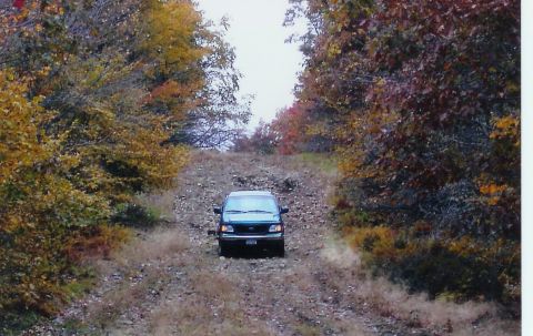 2001 Ford F-150 - A dark pickup truck navigates a gravel road surrounded by trees in autumn colors.