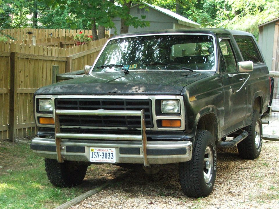 Black two-door Ford Bronco with chrome grille and brush guard parked on gravel by a wood fence