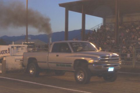 1994 Ford F-150 - A Dodge Ram truck competes in a pull event, kicking up dust and smoke under a stadium roof.