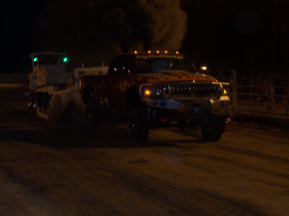 Red pickup truck with flames and "BigBadDodge" text, towing a trailer, emitting black smoke at night