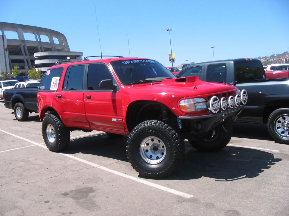 Lifted red SUV with off-road tires, hood scoop, and multiple lights on front bumper. Another truck is parked behind it
