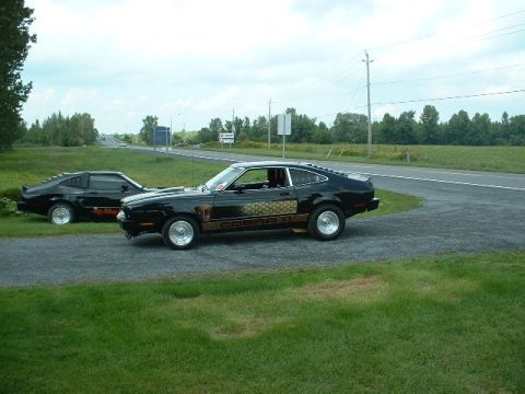 1978 Ford Mustang II - Two black Ford Mustang II Cobras are parked side-by-side on a gravel lot next to a road.
