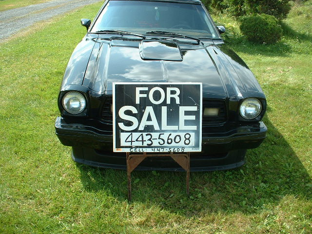 Black 1978 Ford Mustang II, front view, with a 'For Sale' sign on a stand in front