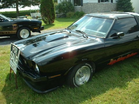1978 Ford Mustang II - A black 1978 Ford Mustang II coupe is parked on a lawn, with a 'For Sale' sign in front and another black car visible behind it.