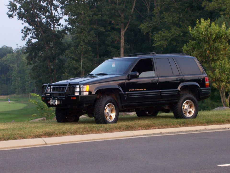 Black 1996 Jeep Grand Cherokee with aftermarket bull bar and gold wheels parked on a grassy incline