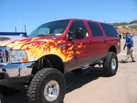 1986 Ford Bronco - This lifted SUV features custom flame graphics and large off-road tires under a clear blue sky.
