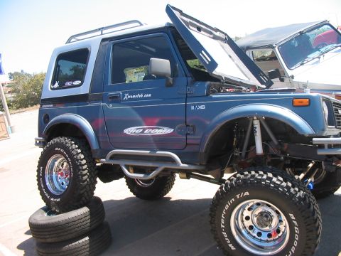 1986 Ford Bronco - A modified blue SUV sits on stacked tires, its hood and windshield open to reveal the engine bay.