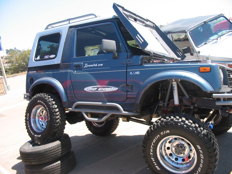 Blue off-road SUV with lifted suspension, hood open, stacked tires under front wheel