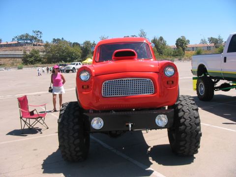 1986 Ford Bronco - A bright red custom truck with oversized tires sits in a parking lot under a clear blue sky.