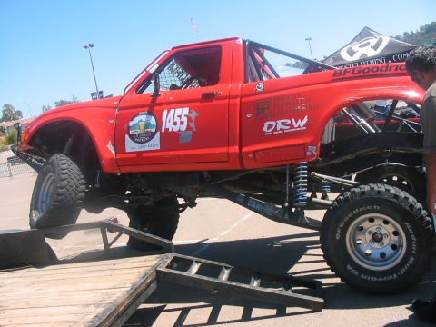 1986 Ford Bronco - A heavily modified red pickup truck with off-road tires is positioned on a ramp, ready for an event.