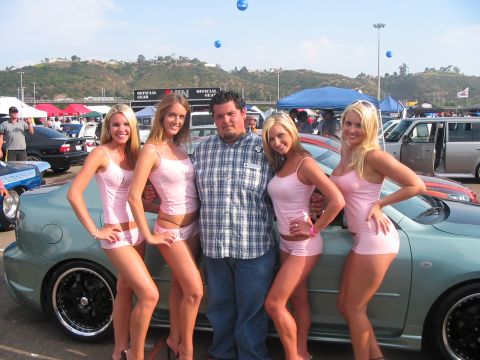 1986 Ford Bronco - A man poses with four women in pink outfits at an outdoor car show, standing in front of a light green/teal compact car with black multi-sp…