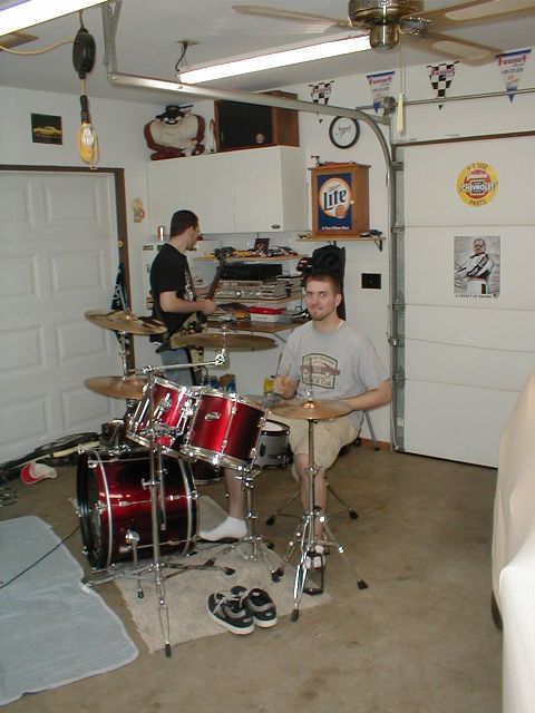 1986 Ford Bronco - Two men are in a garage, one playing a red drum set, with a car partially covered on the right side.