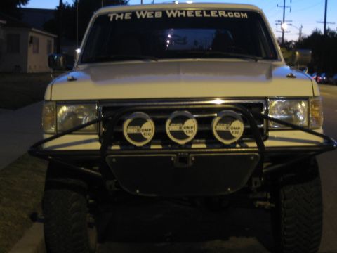1990 Ford Bronco - Front view of a light-colored Ford SUV with a tube bumper and three off-road lights, its windshield reading 'The Web Wheeler.com' at dusk.