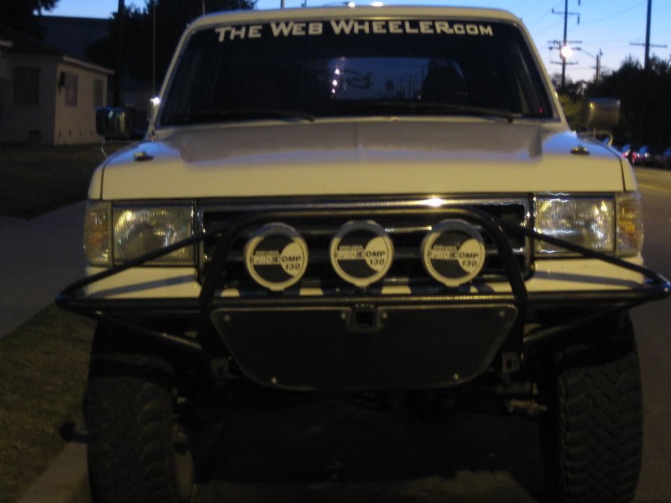 Front view of light-colored Ford SUV with tube bumper, three off-road lights, and 'The Web Wheeler.com' windshield banner